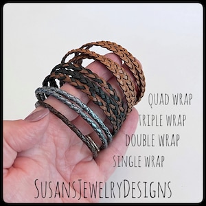 May include: A hand holding a stack of braided leather bracelets in various colors and widths, including brown, black, and silver. The bracelets are labeled "Quad Wrap", "Triple Wrap", "Double Wrap", and "Single Wrap".