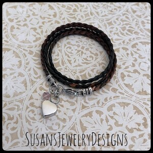 May include: A braided leather bracelet with a silver heart charm and a small pink crystal. The bracelet is brown and black and has a silver clasp.