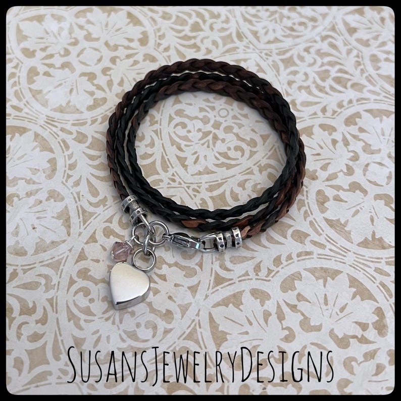 May include: A braided leather bracelet with a silver heart charm and a small pink crystal. The bracelet is brown and black and has a silver clasp.
