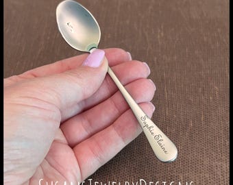 May include: A silver spoon with a rounded bowl and a slender handle. The handle is engraved with the name "Sophie Elaine" in a cursive font. The spoon is held against a brown background.
