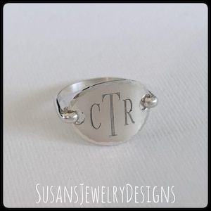 May include: A silver-toned ring featuring a smooth, oval-shaped face plate. The center of the oval is engraved with the letters CTR in a serif font. The band attaches to the sides of the oval plate with small, rounded metal loops on each side.