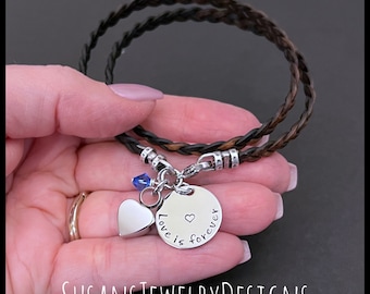 May include: A braided dark brown leather bracelet featuring a silver-toned circular charm engraved with the text "Love is forever" and a small heart symbol. The bracelet also includes a plain silver heart charm and a small blue crystal bead.