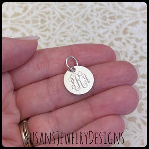 May include: A silver charm with a monogram engraved on a round disc. The charm has a loop for attaching to a necklace or bracelet.