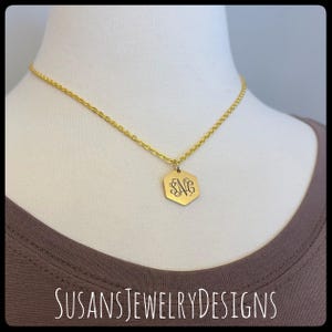 May include: A gold-colored necklace with a hexagonal pendant. The pendant features an engraved monogram. The necklace is displayed on a mannequin. The text "SUSANSJEWELRYDESIGNS" is at the bottom of the image.