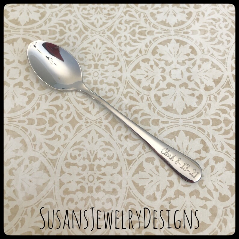 May include: A silver teaspoon with the inscription "Clark 8-18-21" engraved on the handle.  The spoon is on a patterned background.