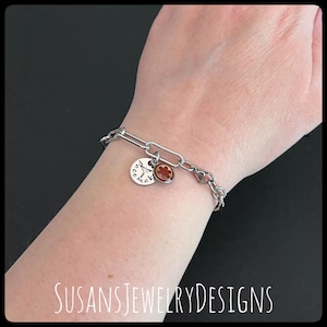 May include: A silver-toned bracelet with a chain link design. The bracelet features two charms: a round charm with the words "peace maker" and a small, reddish-brown charm. The bracelet is worn on a wrist.