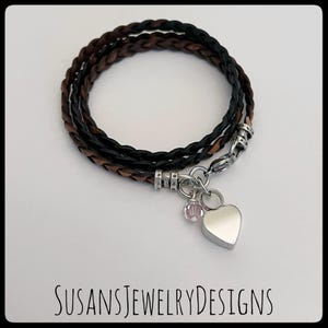 May include: A braided leather bracelet with a silver heart charm and a small pink crystal bead. The bracelet is brown and black.