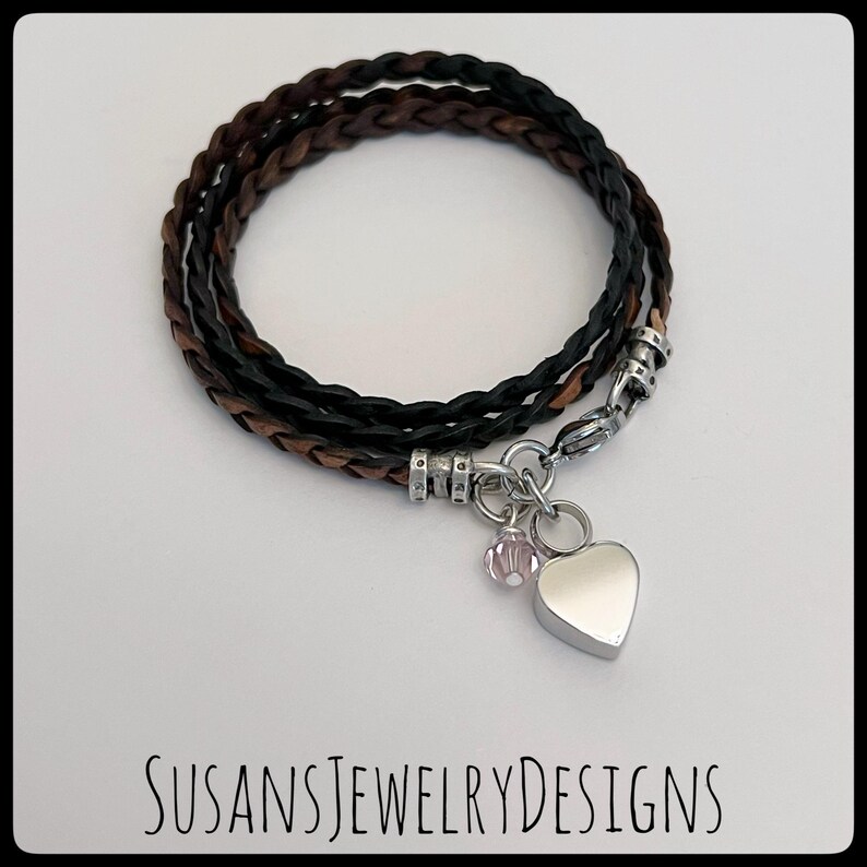 May include: A braided leather bracelet with a silver heart charm and a small pink crystal bead. The bracelet is brown and black.