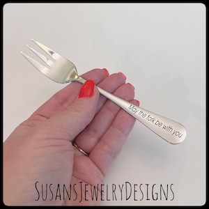 May include: A silver fork with three tines, held in a hand with red nail polish. The fork's handle is engraved with the phrase "May the fork be with you". The image is set against a white background.