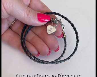 May include: A black leather braided bracelet with a silver heart charm engraved with the letter "M".