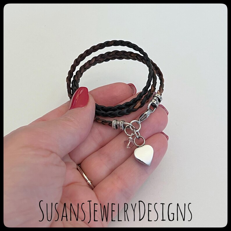 May include: A brown and black braided leather bracelet with a silver heart charm. The bracelet is shown on a hand with red nail polish.