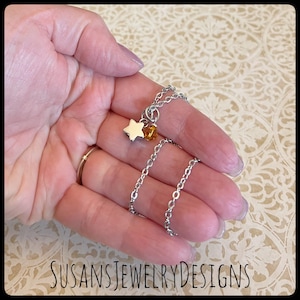 May include: A silver chain necklace with a small silver star charm and a small amber-colored bead charm. The necklace is on a hand.