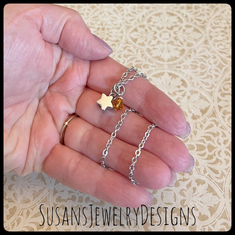 May include: A silver chain necklace with a small silver star charm and a small amber-colored bead charm. The necklace is on a hand.