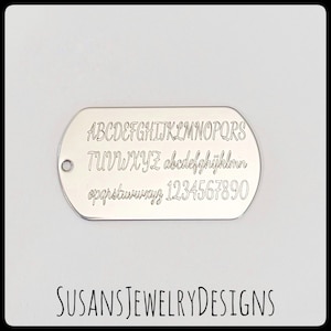 May include: Silver dog tag with a font chart engraved on it. The chart includes uppercase and lowercase letters, numbers, and punctuation. The text "SUSANS JEWELRY DESIGNS" is printed below the dog tag.