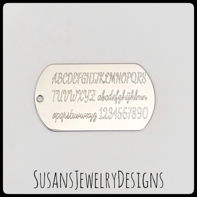May include: Silver dog tag with a font chart engraved on it. The chart includes uppercase and lowercase letters, numbers, and punctuation. The text "SUSANS JEWELRY DESIGNS" is printed below the dog tag.