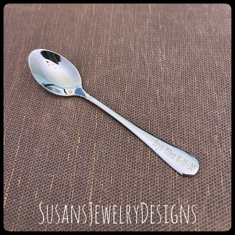 Puede incluir: Una cuchara plateada brillante con un cuenco redondeado y un mango delgado. El mango est&aacute; grabado con el nombre "Bryn Ellen 8-11-20". La cuchara se muestra sobre una superficie marr&oacute;n texturizada. El texto "SUSANSJEWELRYDESIGNS" est&aacute; en la parte inferior.