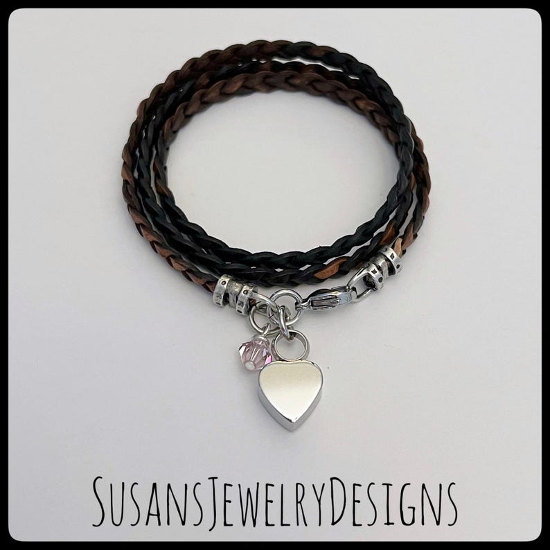 May include: A black and brown braided leather bracelet with a silver heart charm and a small pink crystal. SUSANSJEWELRYDESIGNS is printed on the white background.