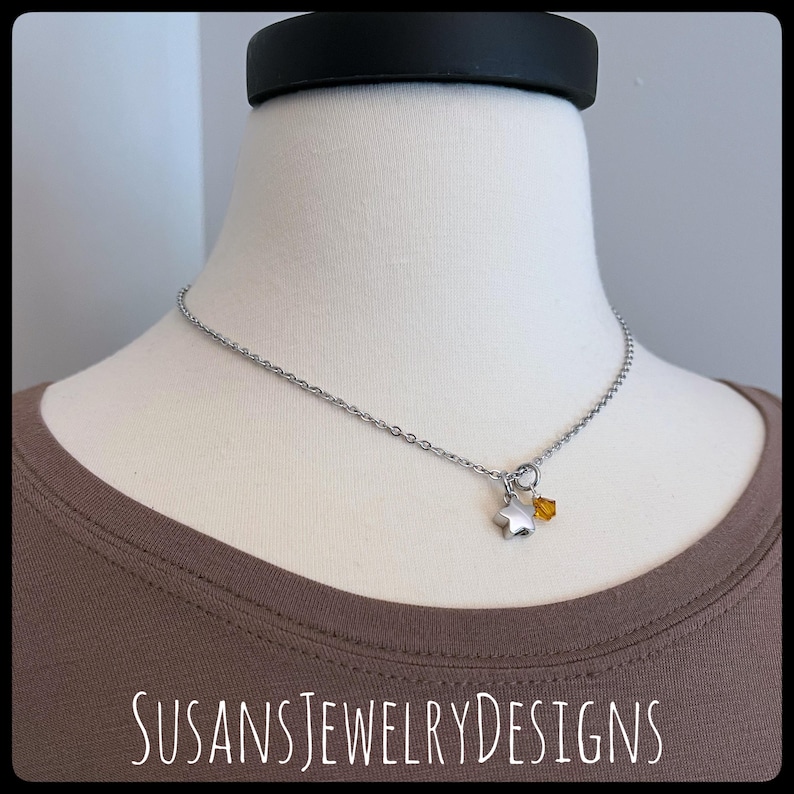 May include: A silver chain necklace with a small silver star charm and a single amber crystal pendant. The necklace is on a brown shirt.