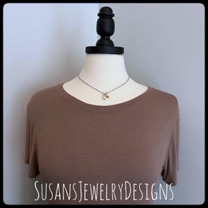 May include: A silver necklace with a small star charm and a small yellow gemstone charm. The necklace is on a brown shirt mannequin.