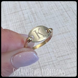 May include: A gold-colored ring with an oval-shaped face engraved with the letter "K". The ring has a delicate band and small ball accents on each side of the oval. The ring is held up by a person's hand.