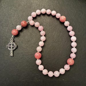 May include: A rosary featuring a series of smooth, round light pink beads interspersed with larger, darker rose-colored beads. A silver-toned metal Celtic cross pendant with intricate knotwork detailing is attached to one end of the bead strand.