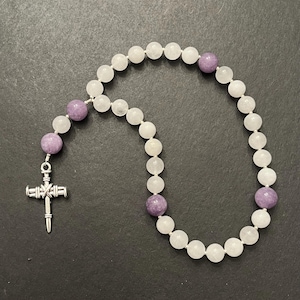 May include: A white and purple beaded prayer mala with a silver cross charm. The mala has 108 beads, with 3 purple beads and the rest white.