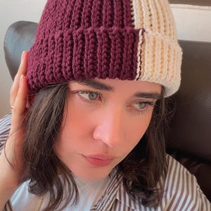 May include: A two-toned knit beanie featuring a ribbed texture. The hat is divided vertically into two distinct sections, with one side in a deep burgundy color and the other side in a light cream color, finished with a folded brim.