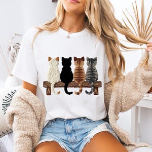 Könnte beinhalten: Weißes T-Shirt mit vier Katzen auf einer Holzbank. Die Katzen sind in den Farben Creme, Schwarz, braun getigert und grau getigert dargestellt. Das T-Shirt hat einen Rundhalsausschnitt.