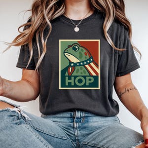 Puede incluir: Camiseta gris oscuro con un gráfico de rana en un estilo de cartel patriótico. La rana lleva un collar con estrellas y una capa roja, blanca y azul. La palabra "HOP" está impresa debajo.