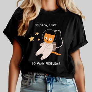 May include: A black t-shirt with a graphic of an orange cat wearing an astronaut suit and the text "Houston, I have so many problems".