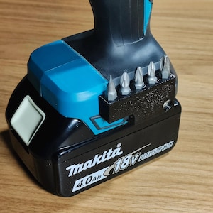 May include: A black and teal Makita 18V cordless drill battery with a set of drill bits. The battery pack has a white square button and the Makita logo. The drill bits are silver and are held in a black holder.