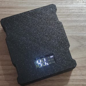 May include: A black, textured power bank with a digital display showing the battery percentage. The power bank has a rectangular shape with a slightly rugged design. The display shows "IN", "OUT", "5V 2.4A", and "QC +" text.