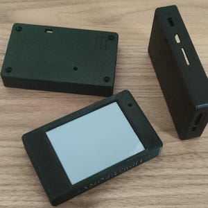Könnte beinhalten: Drei schwarze rechteckige Gehäuse für elektronische Geräte. Eines hat einen Bildschirm, ein anderes eine Reihe von Löchern, und das dritte ist eine einfache Box. Alle sind aus mattem, schwarzem Material.