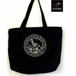 May include: Black canvas tote bag with a circular design featuring a bird on a branch with flowers. The design is made of iridescent materials and is surrounded by a gold geometric pattern.