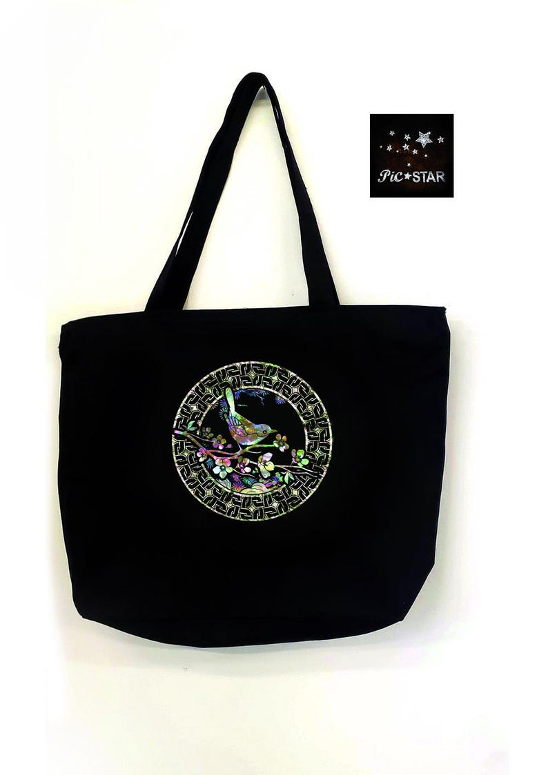 May include: Black canvas tote bag with a circular design featuring a bird on a branch with flowers. The design is made of iridescent materials and is surrounded by a gold geometric pattern.