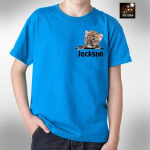 May include: A blue t-shirt featuring a graphic of a leopard cub peeking over the top of a pocket. The name "Jackson" is printed below the graphic.