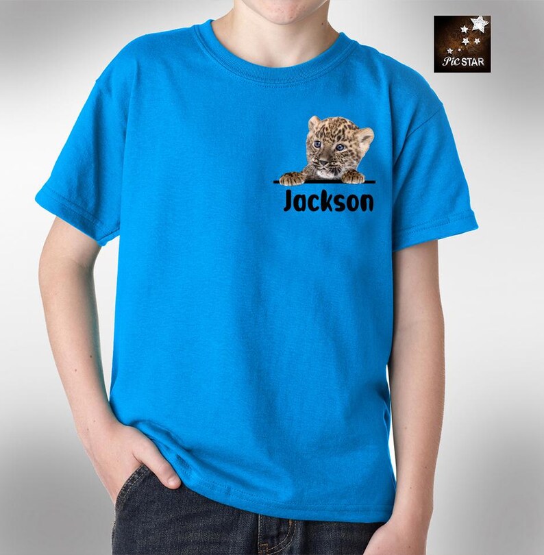 May include: A blue t-shirt featuring a graphic of a leopard cub peeking over the top of a pocket. The name "Jackson" is printed below the graphic.