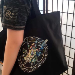May include: Black tote bag with a colorful floral and hummingbird design on the front. The design is surrounded by a geometric pattern.