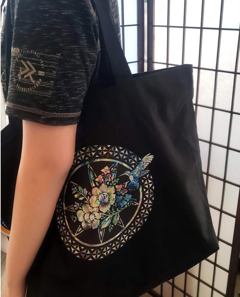 May include: Black tote bag with a colorful floral and hummingbird design on the front. The design is surrounded by a geometric pattern.