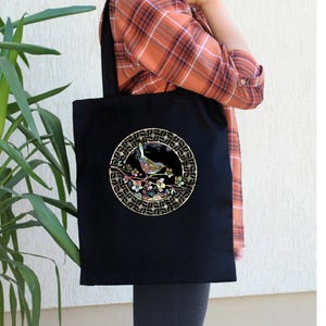 May include: Black tote bag with a circular design featuring a bird perched on a branch with pink flowers. The design is surrounded by a gold geometric pattern.