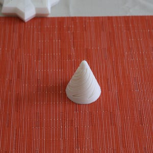 以下が含まれることがあります： 赤い織りマットの上に、らせん模様の白い円錐形の物体が置かれています。この物体は3Dプリントされた素材で作られているようです。背景には、白い星型の物体が部分的に見えています。