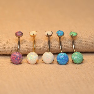 Pode incluir: Seis anéis de umbigo com pedras de opala de diferentes cores. Os anéis são feitos de aço cirúrgico e têm um acabamento dourado.