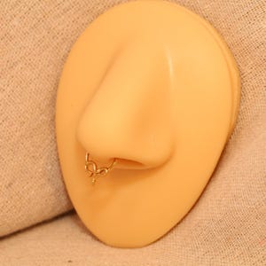 Op de afbeelding: Een goudkleurige septumring met een decoratief, symmetrisch ontwerp met gebogen lijnen en een centraal punt, geplaatst in het neusgat van een beige neusmodel. Het sieraad heeft een gepolijste metalen afwerking en een cirkelvormige ring.