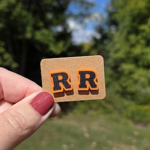 May include: A small, rectangular craft paper tag with the letters "RR" in black with orange outlines. The tag is held up against a backdrop of green foliage and a blue sky.