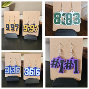 May include: Earrings with sports jersey-style numbers. The earrings are displayed on small cardboard cards. The numbers include 997 in yellow and navy, 83 in green and white, 366 in blue and white, and #1 in purple and black.