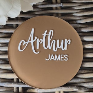 May include: A round, brown acrylic nameplate with the name "Arthur James" written in white cursive lettering.