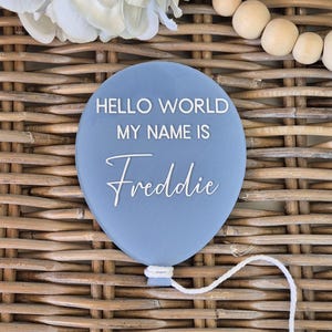 May include: A pale blue balloon-shaped sign with the text "HELLO WORLD MY NAME IS Freddie" in white. The sign has a white string attached and is set against a woven background. Decorative elements include white flowers and wooden beads.