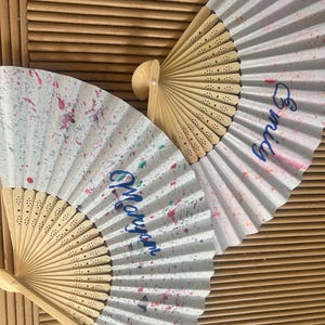 Peut inclure: Deux éventails pliants blancs avec un motif de peinture éclaboussée. Un éventail dit "Morgan" et l'autre dit "Emily".