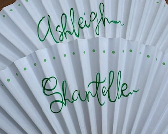 May include: White paper hand fans with green polka dots and handwritten names "Ashleigh" and "Shantelle". The fan has a wooden handle with decorative cutouts.