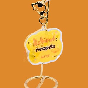 May include: A gold-colored keychain with a yellow cloud-shaped charm. The charm has the words "Retired neopets user" in orange script, with star and jelly-like graphics. The keychain is on a gold stand against an orange background.